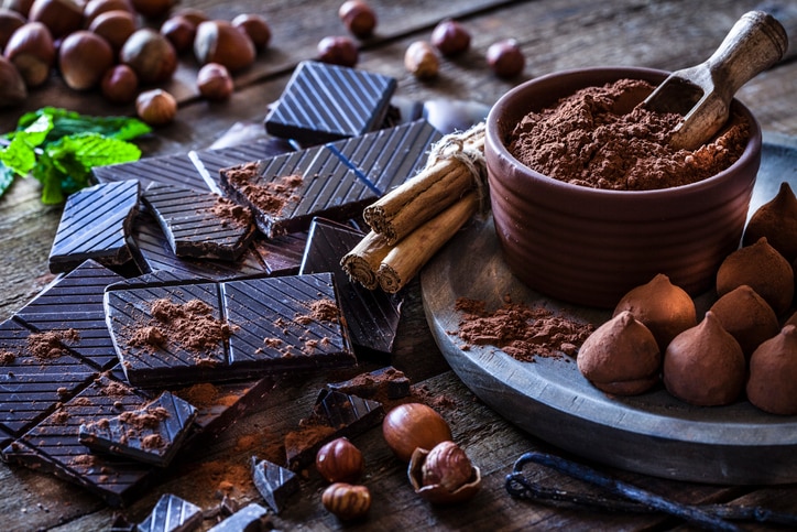 Rustic wooden table filled with ingredient for preparing homemade chocolate truffles. The composition includes dark chocolate truffles, chocolate bars, cocoa powder, cinnamon sticks, vanilla beans, mint leaves and hazelnuts. Predominant color is brown.