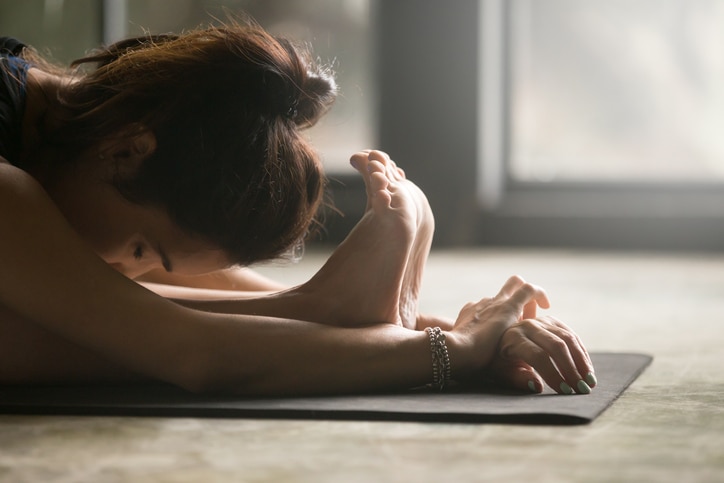 Young attractive woman practicing yoga at home, sitting in paschimottanasana exercise, Seated forward bend pose, model working out, indoor close up image, studio floor background