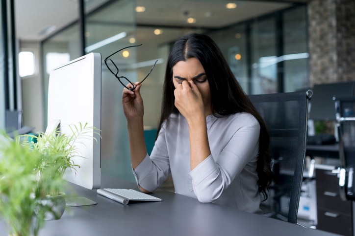 Portrait of depressed young Latin-American businesswoman sitting at computer in office holding glasses and rubbing eyes. Overworking concept