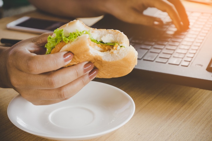 business woman eating junk food burger while working on computer laptop at office desk, unhealthy lifestyle concept