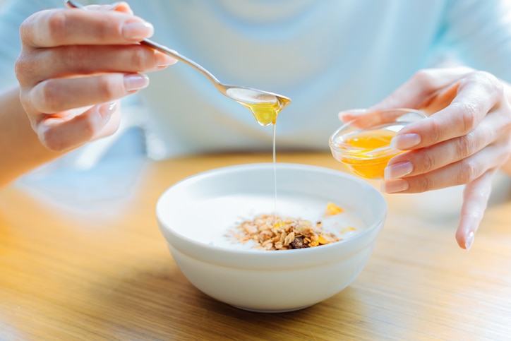 Delicious breakfast. The focus being on the delicate hands of a female adding honey to the bowl of oatmeal with milk standing on the table