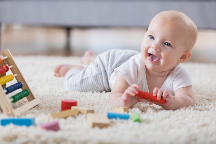 A cute baby lays on her tummy on a rug in a living room and sings with an open mouthed smile.  She is holding one of several wooden blocks.  There is an abacus in the foreground.