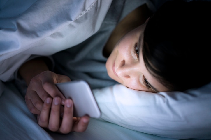 Woman using her phone under blanket in bed at night.