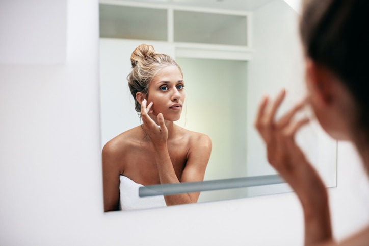 Beautiful young woman standing in the bathroom. Female looking into the mirror and touching her face skin.