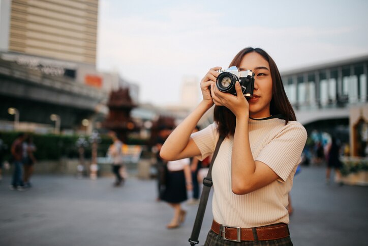 Vintage colored portrait of a young Asian woman traveler, relaxing at the busy Siam station of the Skytrain, next to popular shopping malls and restaurants. She is wearing casual clothing, taking photos with her beautiful analog film camera with a fixed, large aperture, prime lens.