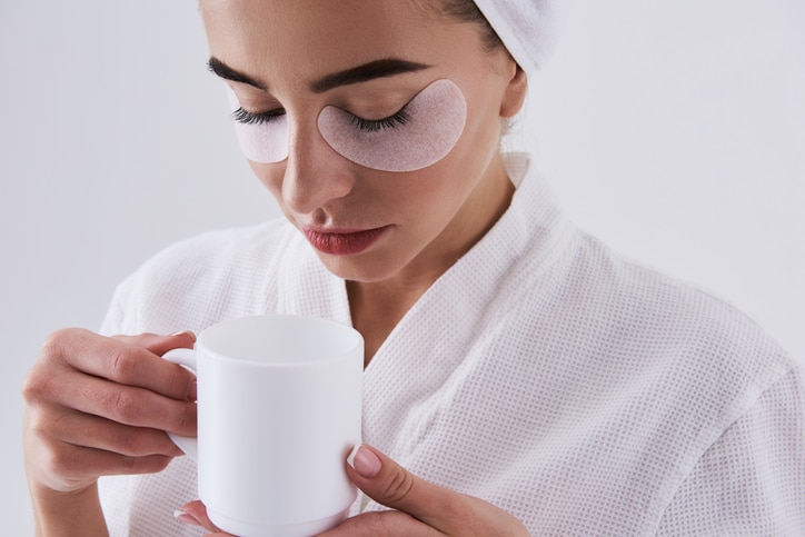 Close up portrait of beautiful girl with masks on lower eyelids holding cup of coffee. Isolated on white background