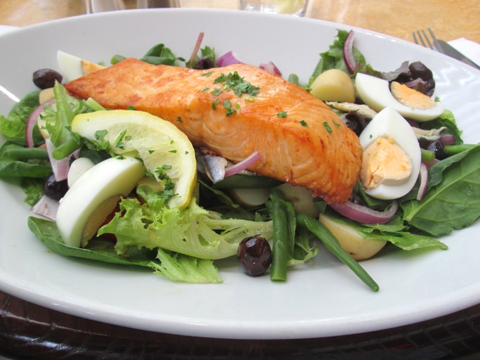 Photo showing a meal of pan fried salmon fillet, served on a dressed salad of lettuce leaves with sliced boiled eggs, red onion rings, chives, green beans / French beans, new potatoes, black olives and a slice of lemon.  This salmon nicoise meal was served as lunch at a restaurant, being pictured on a white plate.