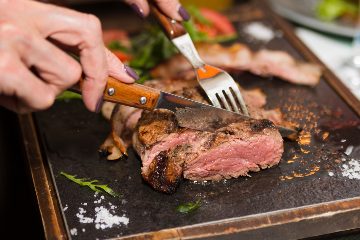 Woman hand holding knife and fork cutting grilled beef steak on stoned plate. Selective focus