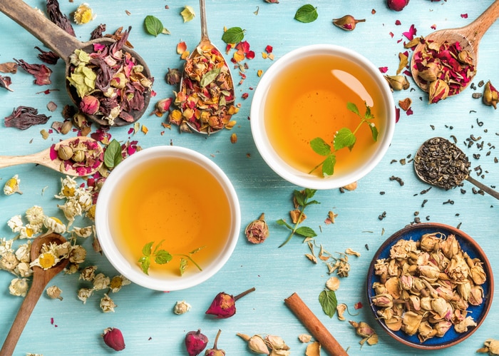 Two cups of healthy herbal tea with mint, cinnamon, dried rose and camomile flowers in spoons over blue background, top view