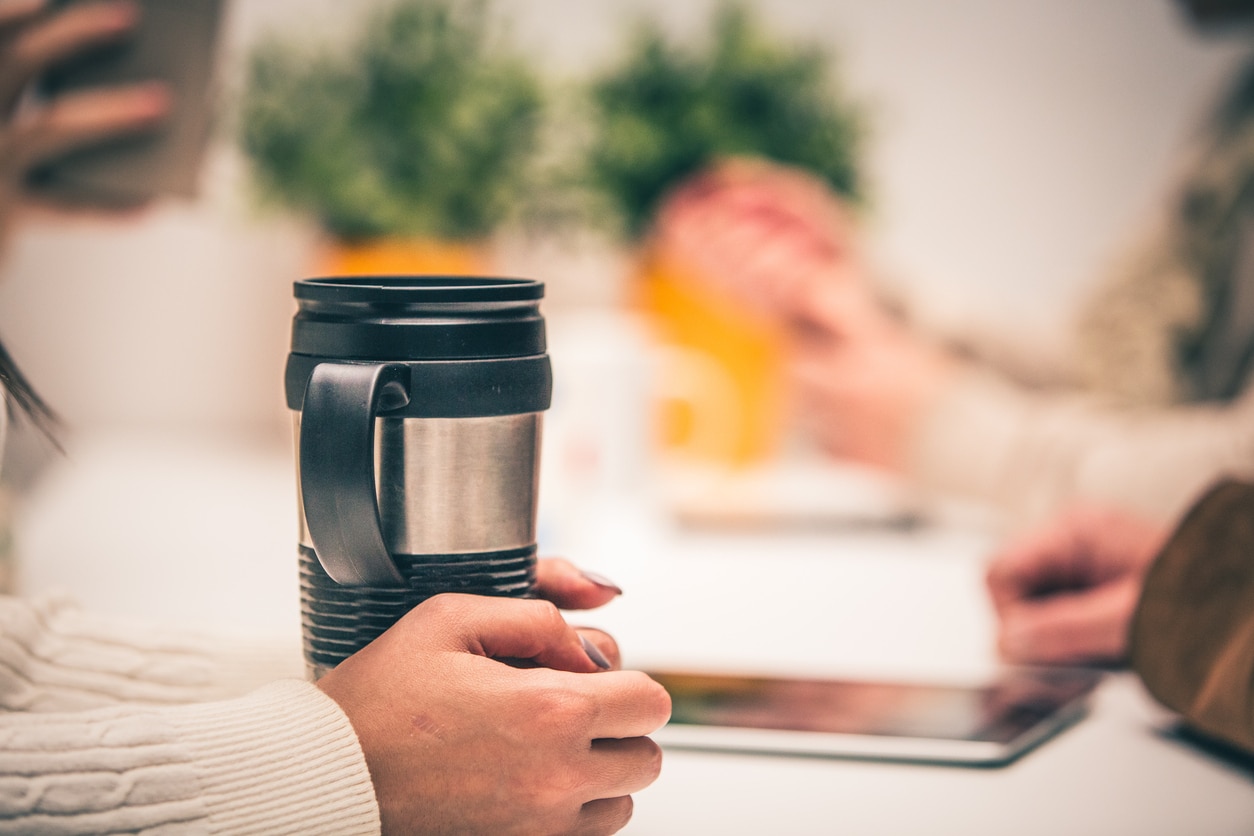 Close-up shot of female hands around travel mug placed on the office desk.