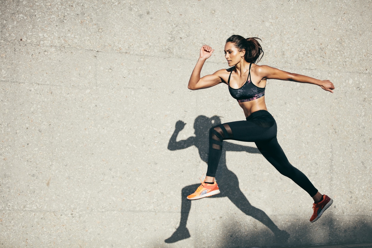 Young woman with fit body jumping and running against grey background. Female model in sportswear exercising outdoors.