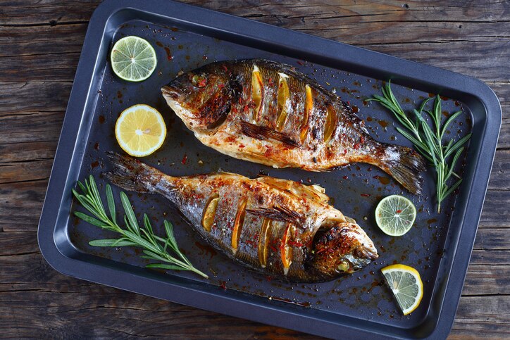 delicious roast dorado or sea bream fish with lemon and orange slices, spices, and rosemary on baking sheets on wooden table, view from above