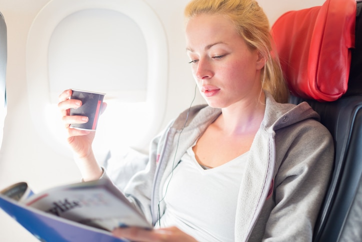 Woman reading magazine and drinking coffeeon airplane. Female traveler reading seated in passanger cabin. Sun shining trough airplane window.