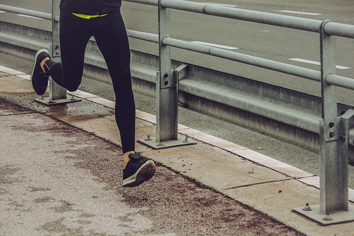 Young fit person running and listening to music in an urban environment. Black sport clothing - sport shoes, running tights, and a jacket.