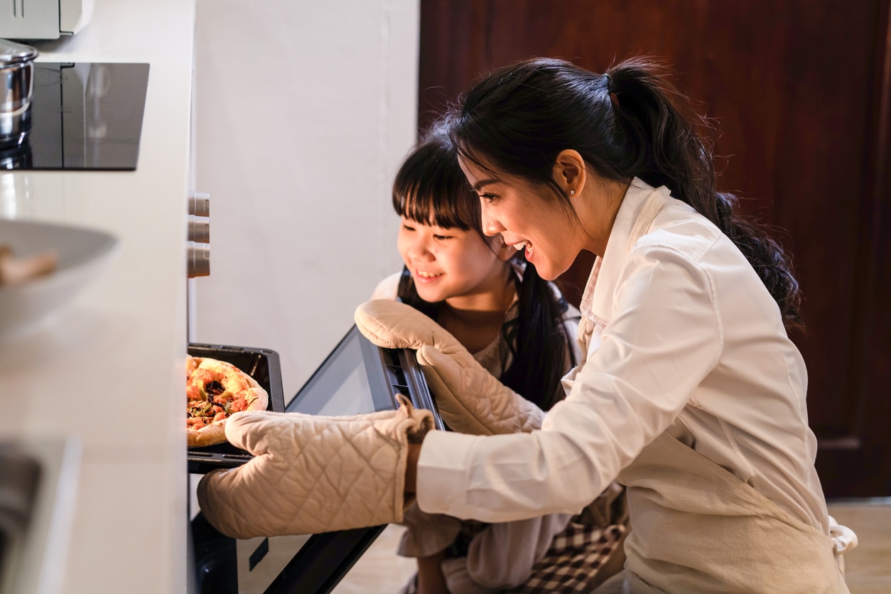 Asian young mother and daughter making pizza at home. Woman open the oven and bring the food out from machine. Little girl looking at meal and smell it with smile face, enjoy famiy activity together.