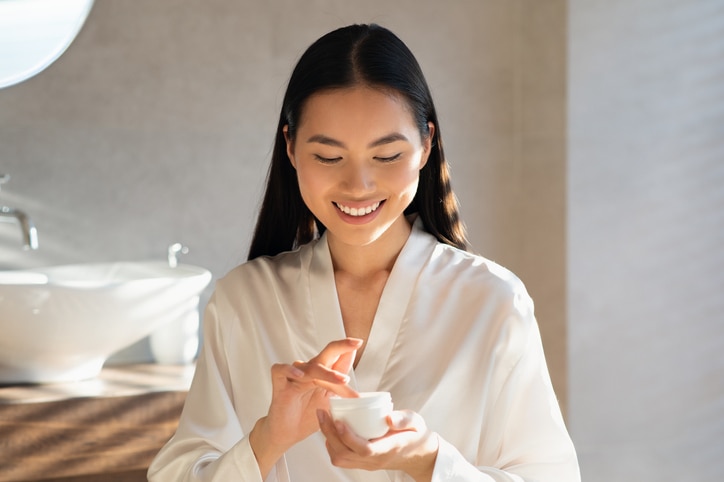 Smiling attractive young asian lady in white bathrobe holding jar with beauty product, MBTI 16 Personalities – The best skincare gift for ISTP